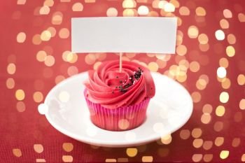 food, baking and pastry concept - close up of cupcake or muffin on plate with red buttercream frosting and blank nametag over festive lights. close up of cupcake with red buttercream frosting