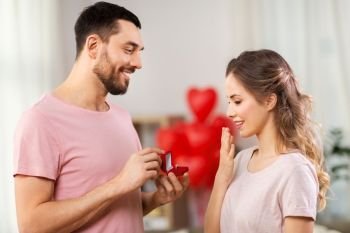love, couple, proposal and people concept - happy man giving diamond engagement ring in little red box to woman at home. man giving woman engagement ring on valentines day