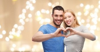 love and family concept - smiling couple showing hand heart gesture over beige background with festive lights. couple showing hand heart over festive lights