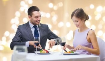 people and leisure concept - smiling couple eating appetizers at restaurant over festive lights on beige background. smiling couple eating appetizers at restaurant