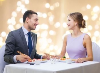 people and food concept - smiling couple eating sushi rolls by chopsticks at restaurant over festive lights on beige background. smiling couple eating sushi rolls at restaurant