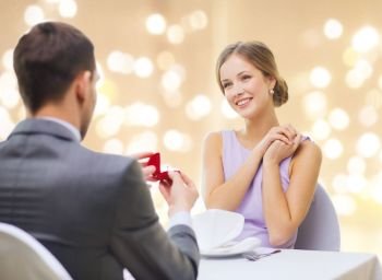 proposal, valentines day and people concept - excited young woman looking at boyfriend giving her engagement ring at restaurant over festive lights on beige background. man giving woman engagement ring at restaurant