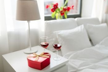 valentines day, love and romantic concept - heart shaped gift box and two glasses of red wine on bedroom nightstand at home. gift and two glasses of wine in bedroom at home