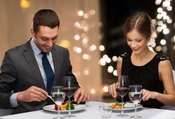 people and leisure concept - smiling couple eating main course with red wine at restaurant over festive lights on background. smiling couple eating main course at restaurant