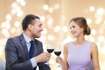 dating, celebration and valentines day concept - smiling young couple clinking glasses of non-alcoholic red wine and looking at each at restaurant other over festive lights on beige background. young couple with glasses of wine at restaurant
