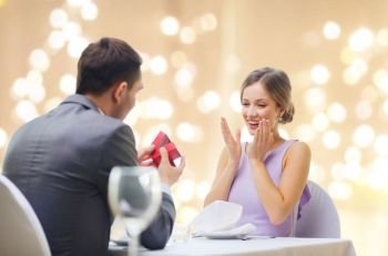 proposal, valentines day and people concept - happy man showing woman present in red box at restaurant over festive lights on beige background. man showing woman present in red box at restaurant