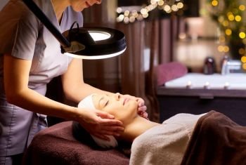 people, beauty, lifestyle and relaxation concept - beautician with magnifying lamp doing face massage to beautiful young woman at spa. beautician doing face massage to woman at spa