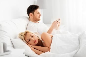 people, technology, internet addiction and cheating concept - man using smartphone while his wife or girlfriend is sleeping in bed. man using smartphone while woman is sleeping