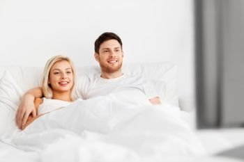 people, leisure and rest concept - happy couple lying in bed at home and watching tv. happy couple lying in bed at home and watching tv