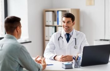 medicine, healthcare and people concept - smiling doctor talking to male patient at medical office in hospital. doctor with laptop and male patient at hospital