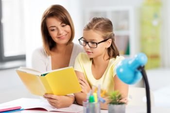 education, family and learning concept - mother and daughter doing homework together and reading book at home. mother and daughter doing homework together