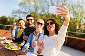 leisure and people concept - happy friends with drinks having dinner party on rooftop in summer and taking selfie by smartphone. happy friends taking selfie at rooftop party