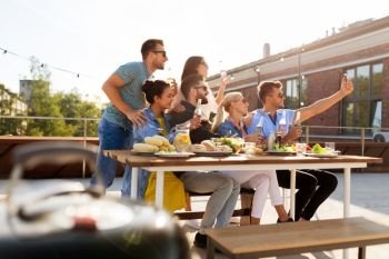 leisure and people concept - happy friends with drinks having barbecue party on rooftop in summer and taking selfie by smartphone. happy friends taking selfie at rooftop party