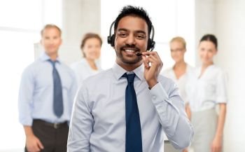 business, customer service and people concept - smiling indian businessman or helpline operator in headset over colleagues on background. indian businessman or helpline operator in headset