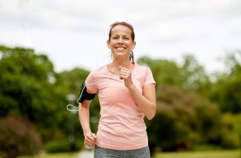 fitness, sport and healthy lifestyle concept - smiling woman with earphones wearing armband for smartphone, jogging at summer park and listening to music. woman with earphones add armband jogging at park
