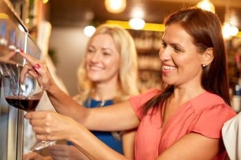 people, alcohol and lifestyle concept - happy women with glass pouring red wine from dispenser at bar or restaurant. happy women pouring wine from dispenser at bar