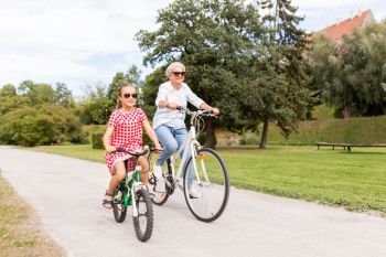 family, leisure and people concept - happy grandmother and granddaughter riding bicycles at summer park. grandmother and granddaughter cycling at park