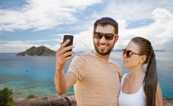 travel, tourism and people concept - happy couple taking selfie by smartphone over tropical beach on seychelles island background. couple taking selfie by smartphone on seychelles