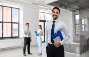 business, realty and people concept - smiling indian businessman or realtor with clipboard over empty office room background. indian businessman or realtor in empty office room