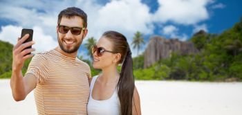 travel, tourism and people concept - happy couple taking selfie by smartphone over tropical beach on seychelles island background. happy couple taking selfie by smartphone on beach