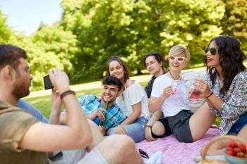 friendship, leisure and technology concept - guy taking picture on smartphone of friends drinking non alcoholic drinks at picnic in summer park. guy taking picture of friends on summer picnic