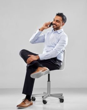 business, communication and people concept - smiling indian businessman sitting on office chair and calling on smartphone over grey background. indian businessman calling on smartphone
