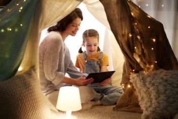 family, hygge and technology concept - happy mother and little daughter with tablet pc computer in kids tent at night at home. family with tablet pc in kids tent at home