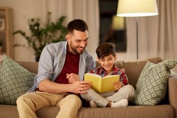 family, childhood, fatherhood, leisure and people concept - happy smiling father and little son reading book on sofa at home in evening. happy father and son reading book sofa at home