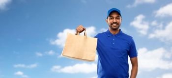 service and people concept - happy indian delivery man food in paper bag in blue uniform over sky and clouds background. happy indian delivery man with food in paper bag