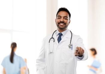 medicine, profession and healthcare concept - smiling indian male doctor in white coat with stethoscope pointing finger to you over hospital background. smiling indian male doctor pointing finger to you