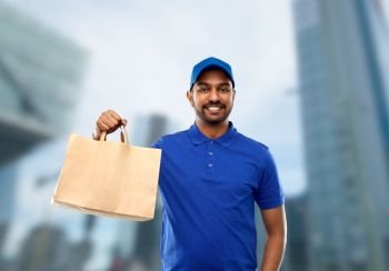 service and people concept - happy indian delivery man food in paper bag in blue uniform over tokyo city skyscrapers background. happy indian delivery man with food in paper bag