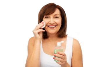 beauty, skin care and old people concept - smiling senior woman cleaning her face with lotion and cotton pad over white background. senior woman cleaning face by lotion on cotton pad