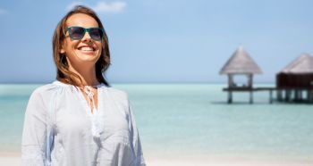 people and leisure concept - happy smiling woman in sunglasses over tropical beach and bungalow on maldives background. happy smiling woman in sunglasses over beach