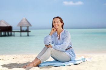 summer, people and leisure concept - happy smiling woman sitting on towel over tropical beach and bungalow on maldives background. happy woman sitting on summer beach