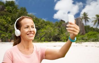 fitness, technology and healthy lifestyle concept - smiling woman in headphones listening to music and taking selfie by smartphone over tropical beach on seychelles island background. woman in phones takes selfie by cellphone on beach