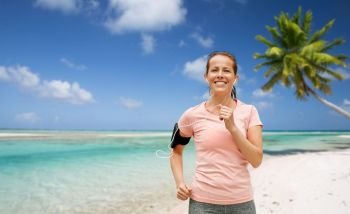 fitness, sport and healthy lifestyle concept - smiling woman with earphones wearing armband for smartphone running and listening to music over tropical beach background in french polynesia. woman with earphones and armband running on beach