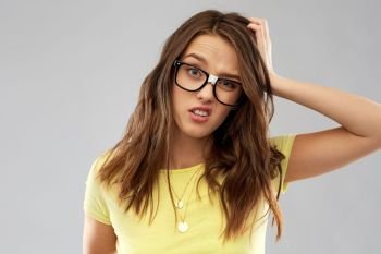 people, emotion and expression concept - confused young woman or teenage girl in yellow t-shirt and glasses over grey background. confused young woman or teenage girl in glasses