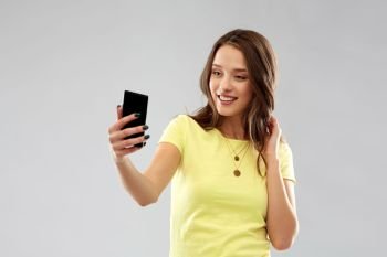 technology and people concept - smiling young woman or teenage girl in blank yellow t-shirt taking selfie by smartphone over grey background. smiling teenage girl taking selfie by smartphone