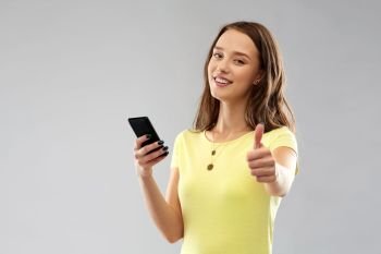 technology and people concept - smiling young woman or teenage girl in blank yellow t-shirt with smartphone showing thumbs up over grey background. teenage girl with smartphone showing thumbs up