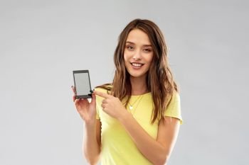 technology and people concept - smiling young woman or teenage girl in yellow t-shirt pointing finger to smartphone blank screen over grey background. young woman or teenage girl holding smartphone