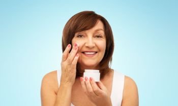 beauty, skincare and cosmetics concept - smiling senior woman applying cream to her face over blue background. smiling senior woman applying cream to her face