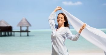 people and leisure concept - happy woman with shawl waving in wind over tropical beach and bungalow on maldives background. happy woman with shawl waving in wind on beach