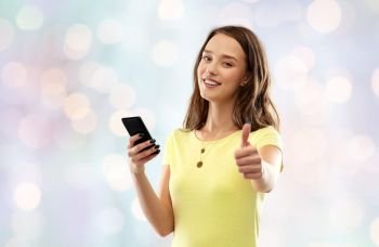 technology and people concept - smiling young woman or teenage girl in blank yellow t-shirt with smartphone showing thumbs up over festive lights background. teenage girl with smartphone showing thumbs up