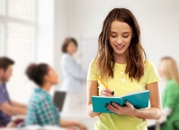high school, education and people concept - smiling young teenage student girl in yellow t-shirt writing to diary or notebook by pencil over classroom and teacher background. teenage student girl writing to notebook at school