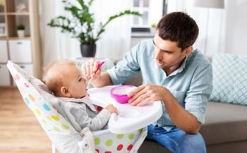 family, food, eating and people concept - happy father feeding little baby daughter sitting in highchair with puree by spoon at home. father feeding happy baby in highchair at home