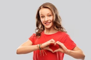 valentine’s day, love and charity concept - smiling teenage girl with long hair in red t-shirt making hand heart gesture over grey background. smiling teenage girl in red making hand heart