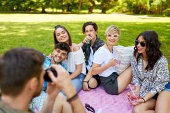 friendship, leisure and technology concept - man with camera photographing his friends drinking non alcoholic drinks at picnic in summer park. friends with drinks photographing at summer picnic