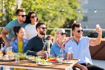leisure and people concept - happy friends with non alcoholic drinks having dinner party on rooftop in summer and taking selfie by smartphone. happy friends taking selfie at rooftop party