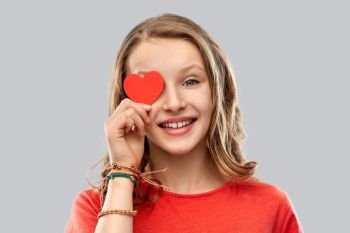 valentine’s day, love and people concept - smiling pretty teenage girl covering one eye with red heart over grey background. smiling teenage girl covering eye with red heart