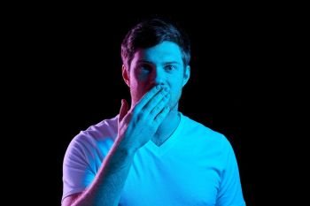 emotion, expression and people concept - portrait of young man in t-shirt covering his mouth by hand over ultra violet neon lights in dark room. man covering his mouth by hand in neon lights